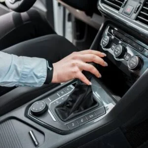 A man wearing a light blue shirt is adjusting the gear shifter in a car's center console. The car's dashboard, including various control buttons and air conditioning dials.