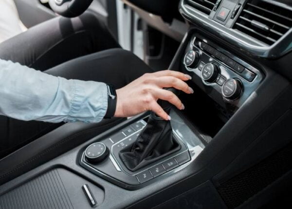 A man wearing a light blue shirt is adjusting the gear shifter in a car's center console. The car's dashboard, including various control buttons and air conditioning dials.