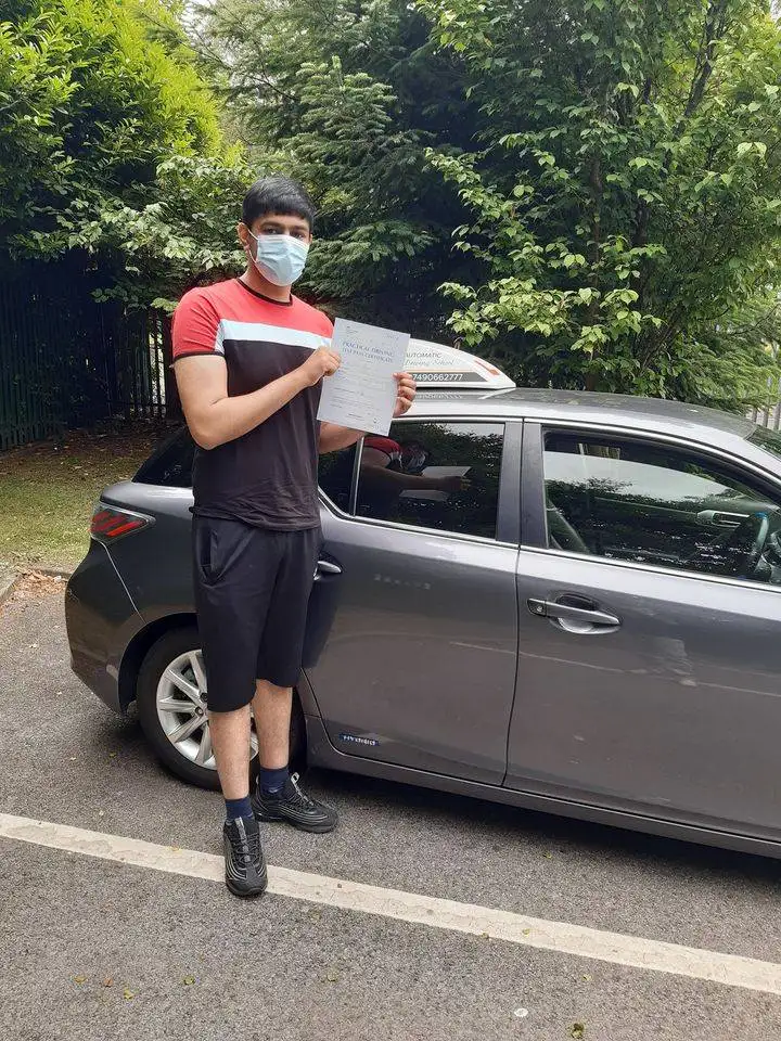 A happy learner standing beside a car, proudly holding a pass test certificate, symbolizing successful driving lessons and test preparation with Shah Driving School in Bolton.