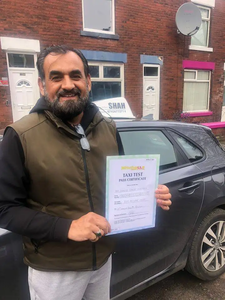 An older man standing proudly next to a car, smiling and holding his driving test pass certificate, celebrating his success with Shah Driving School in Bolton.