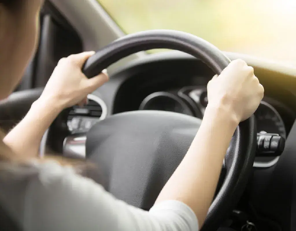 Close-up of a person gripping the steering wheel of a car with both hands. The interior of the car's dashboard and steering column is visible.