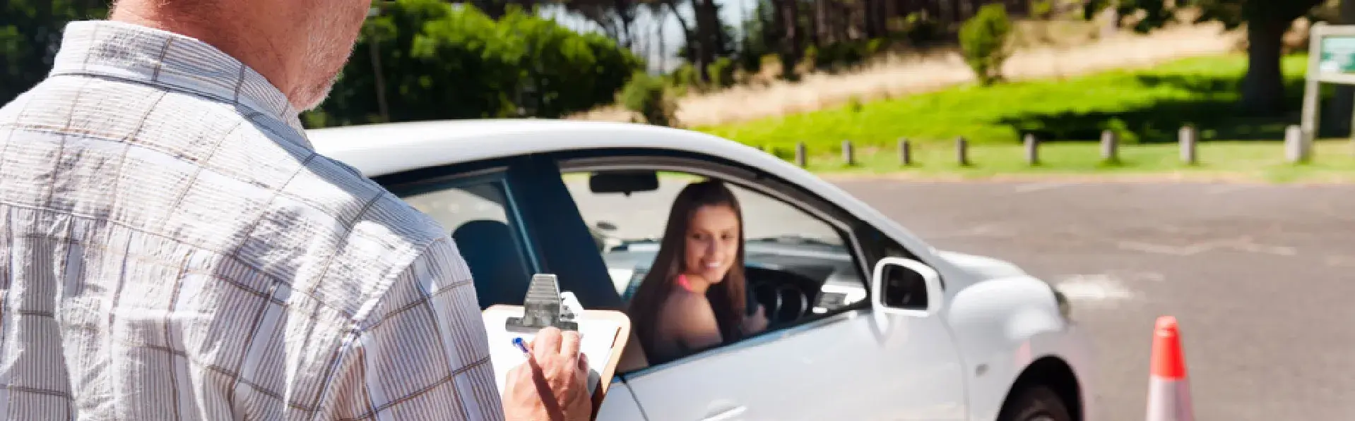 Driving instructor is holding a clipboard and standing outside near an empty parking lot. A woman inside a car is looking at the instructor. Traffic cones are visible in the background.