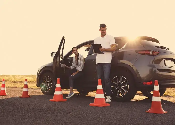 Woman exits a car parked on a road, surrounded by traffic cones. A driving instructor stands beside the car, holding a clipboard.