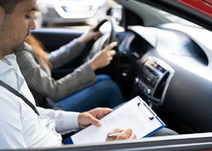 Driving instructor is taking notes on a clipboard while seated in the passenger seat of a car. Another person, seated in the driver's seat, is holding the steering wheel.