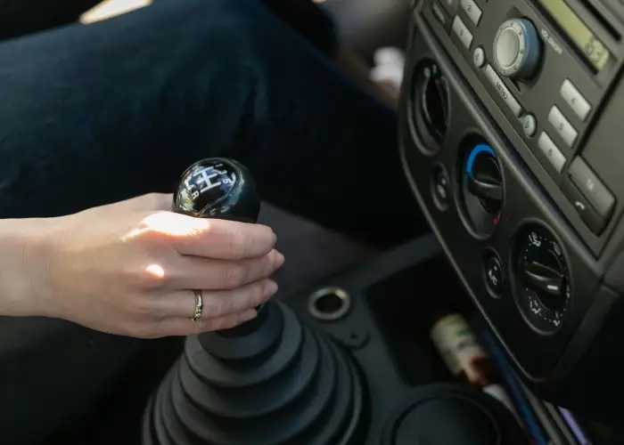 Close-up of a person's hand gripping a manual gear shift in a car. The car interior features a dashboard with heating and cooling controls, a radio, and various buttons and knobs.