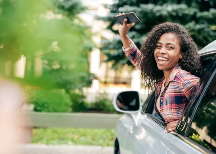 Woman leans out of a car window, holding a smartphone in her raised hand. The background features greenery and trees.