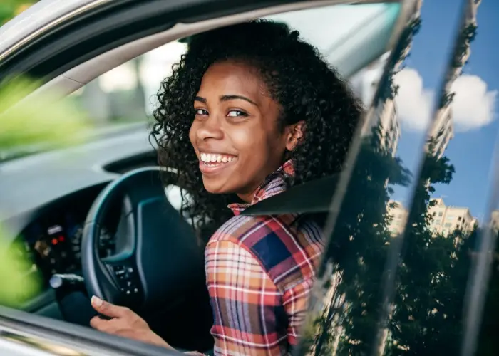 A woman with curly hair, is smiling while looking back from the driver's seat of a car. The window reflects trees and a partly cloudy sky.
