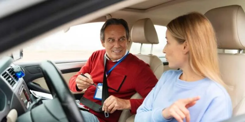 A man seated in the driver's seat of a car, talking to a woman sitting in the passenger seat. The woman, appears to be asking a question. Indicating that the man might be her driving instructor.