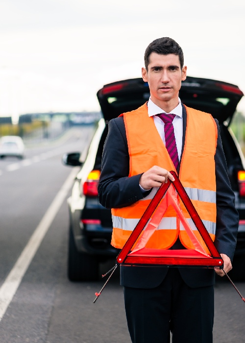 A man in a suit and orange safety vest holds a red reflective warning triangle on a road, exemplifying the everyday conveniences of preparedness.