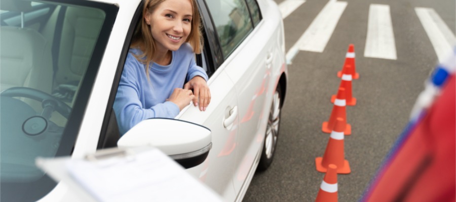 The scene appears to be part of a driving test, A young woman sits in driver's seat of a white car, parked next to orange traffic cones. With a clipboard likely held by a driving instructor visible in the foreground.