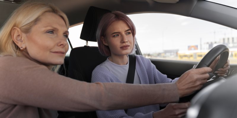 A young woman is learning to drive, holding the steering wheel and looking intently ahead. A female driving instructor sits in the passenger seat, providing guidance.