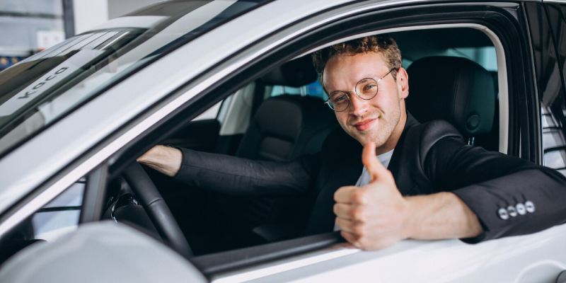 Man sits in the driver's seat of a white car during a driving lesson. He is smiling and giving a thumbs-up with his right hand, which is extended out the open window.