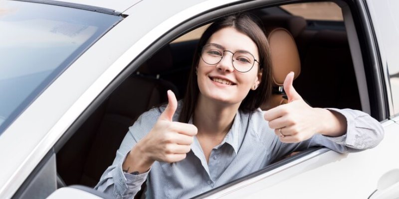 A young woman with glasses, seated in the driver's seat of a car, smiles while giving two thumbs up through the open window. 