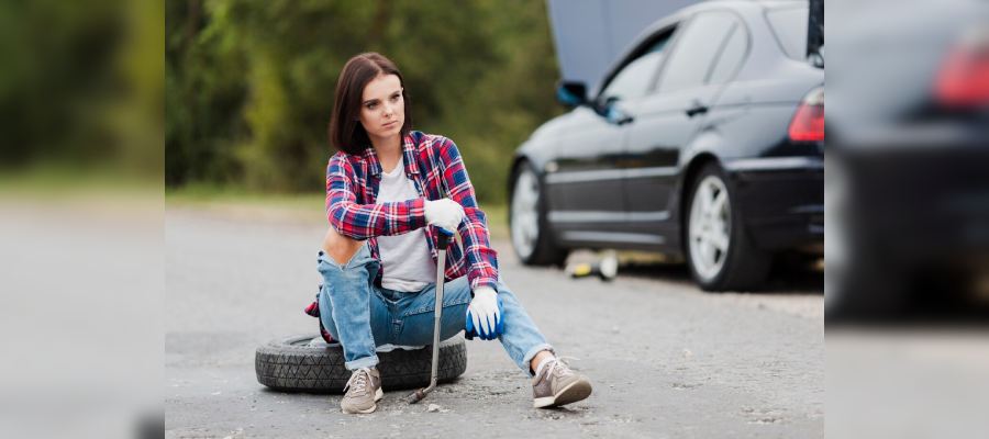 A woman wearing a plaid shirt, jeans sits on the ground next to a tire, leaning against a car jack. She looks tired or frustrated, possibly due to a flat tire or car trouble.