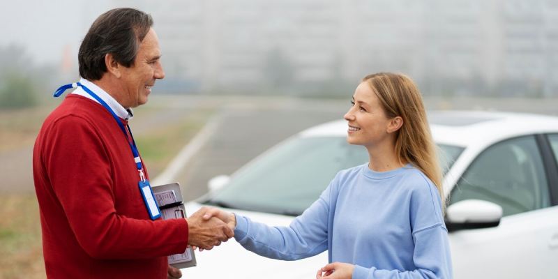 A driving instructor shakes hands with a woman. They are outdoors, standing in front of a white car. The man holds some documents and wears an ID badge around his neck.