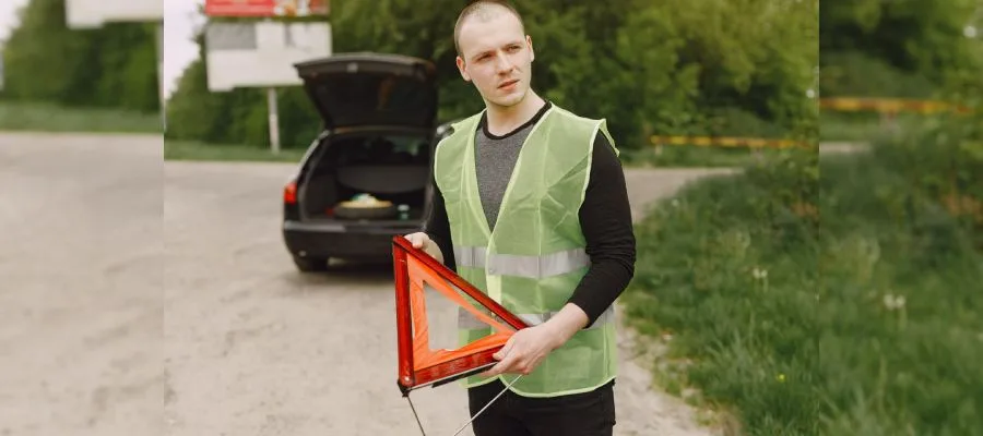 A man wearing a green reflective safety vest is standing roadside, holding a red warning triangle. He appears to be preparing to set up the warning triangle near his car, possibly as part of a driving test.