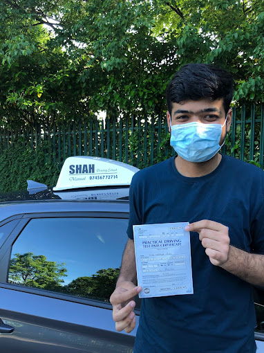 A person wearing a mask standing next to a car and holding a Practical Driving Test Pass Certificate. The car has a "SHAH Driving School" sign on the roof with a phone number.
