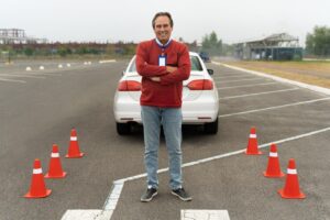 A driving instructor stands confidently in front of a white car in an empty parking lot. Orange traffic cones are arranged in a semi-circle around him.