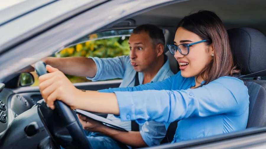 Woman learning to drive in a car, gripping the steering wheel with a focused expression. A driving instructor, is sitting in the passenger seat, and holding onto the dashboard.