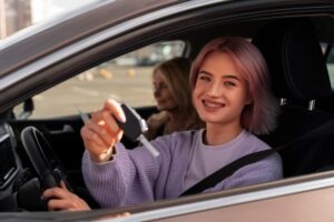 A young person holds up a car key while sitting in the driver's seat of a car. likely taking a driving lesson.