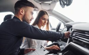 A man, holding a clipboard and appearing to be a driving instructor, is pointing at the car's dashboard while the woman, who is in the driver's seat, listens attentively.