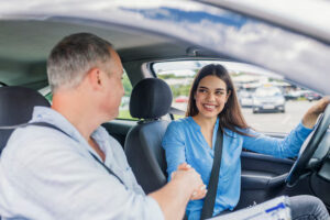 A young woman, seated in a car's driver seat, smiles and shakes hands with her driving instructor in the passenger seat. 