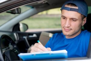 A person wearing a blue shirt and matching cap is seated in a car, holding a pen and writing on a clipboard during a driving lesson.
