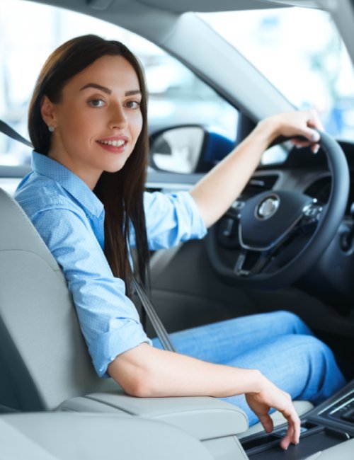 A woman is sitting in the driver's seat of a car, taking an automatic driving lesson. One hand on the steering wheel and the other on the center console.