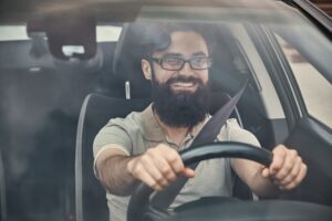 A bearded man wearing glasses and a casual shirt is smiling while driving a car. Holding the steering wheel with both hands, and appears to be enjoying the drive during his driving lesson.driving lesson.