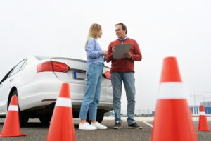 A woman and a man stand in front of a white car during a driving lesson or test. The man is holding a clipboard and appears to be giving instructions or feedback. 