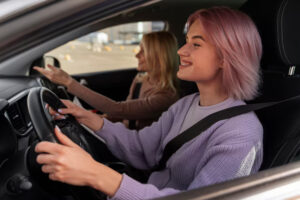 A woman is taking driving lessons smiling and concentrating on the road with her female driving instructor pointing forward, appearing to give directions or guidance.