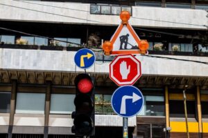 A traffic signal with a red light is paired with several road signs, ideal for beginner drivers to hone their driving skills. The signs include a construction warning at the top, a stop sign with a hand symbol in the middle, and two blue circular signs indicating right turns. 