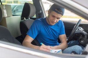 A man sits in the driver's seat writing on a clipboard. The car window is open, and he appears focused on his driving theory test