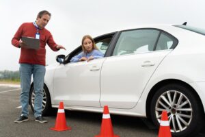 A driving instructor stands beside a white car, holding a clipboard and pointing towards the front of the vehicle. A young woman sits in the driver's seat, looking at the instructor.