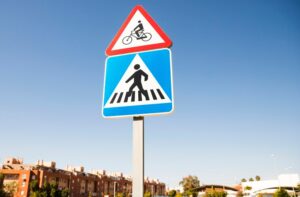 A road sign with a red and white triangle at the top shows a bicycle, indicating a cycle path. Below, a blue and white square sign features a pedestrian on a crosswalk, signaling a pedestrian crossing ahead.