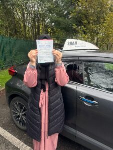 Happy female student holding a driving test pass certificate next to a Shah Driving School car in Bolton, demonstrating the success of our lady driving tutors.