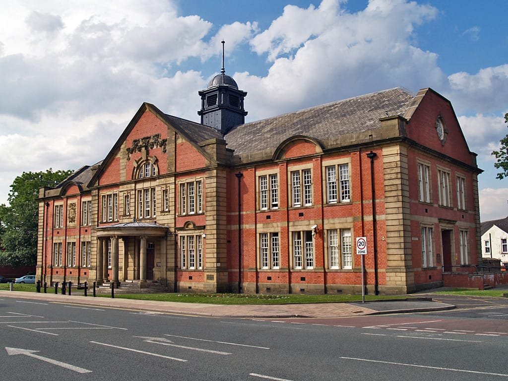 Red brick Edwardian architecture of Farnworth Town Hall with a stone portico, cupola, and multi-paned windows under a partly cloudy sky.