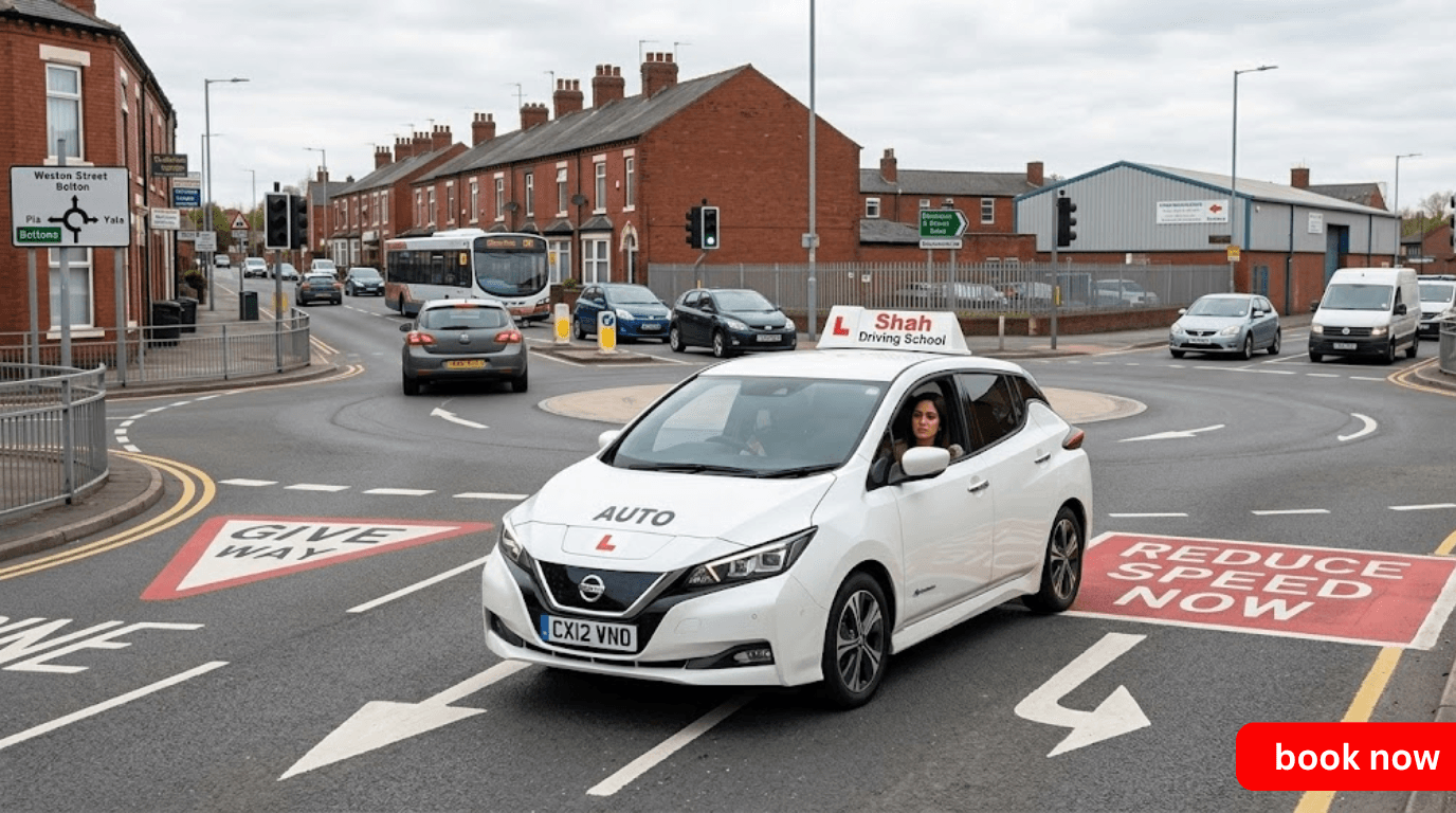 A white automatic Shah Driving School tuition car with an L-plate roof sign navigating a multi-lane roundabout near Weston Street in Bolton, featuring prominent road markings like 'GIVE WAY' and 'REDUCE SPEED NOW'.
