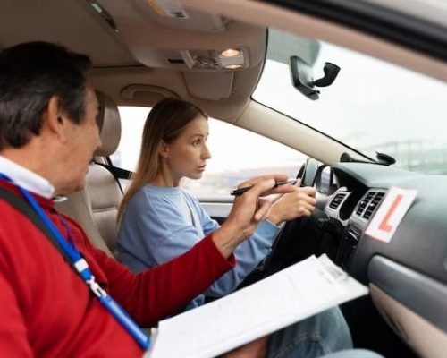 Young woman sits in the driver's seat of a car, holding the steering wheel tightly while listening to a middle-aged driving instructor who is pointing towards the dashboard.