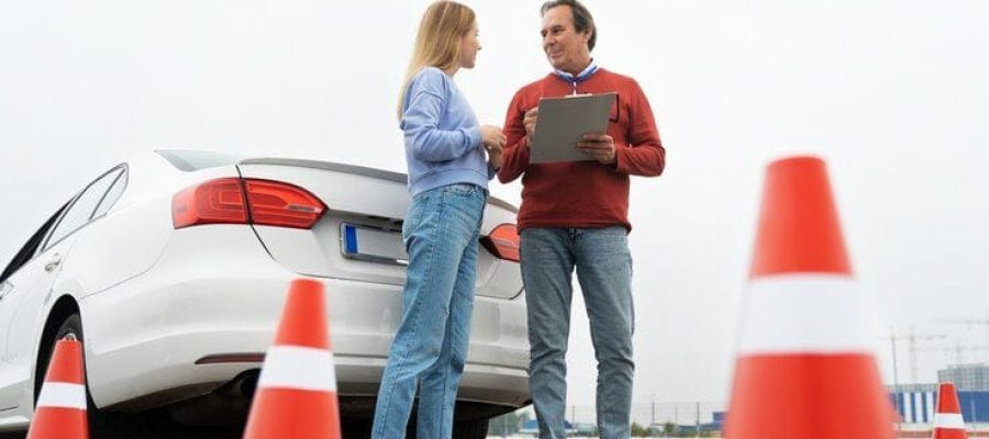 A woman with her driving instructor stand next to a white car with traffic cones around them. They appear to be discussing something, possibly a driving lesson or a driving test.